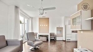 A modern living room with a sofa, a chair, a dining table, and a kitchen area with cabinets and a TV. - Markgraf Apartments (Bad Bellingen)