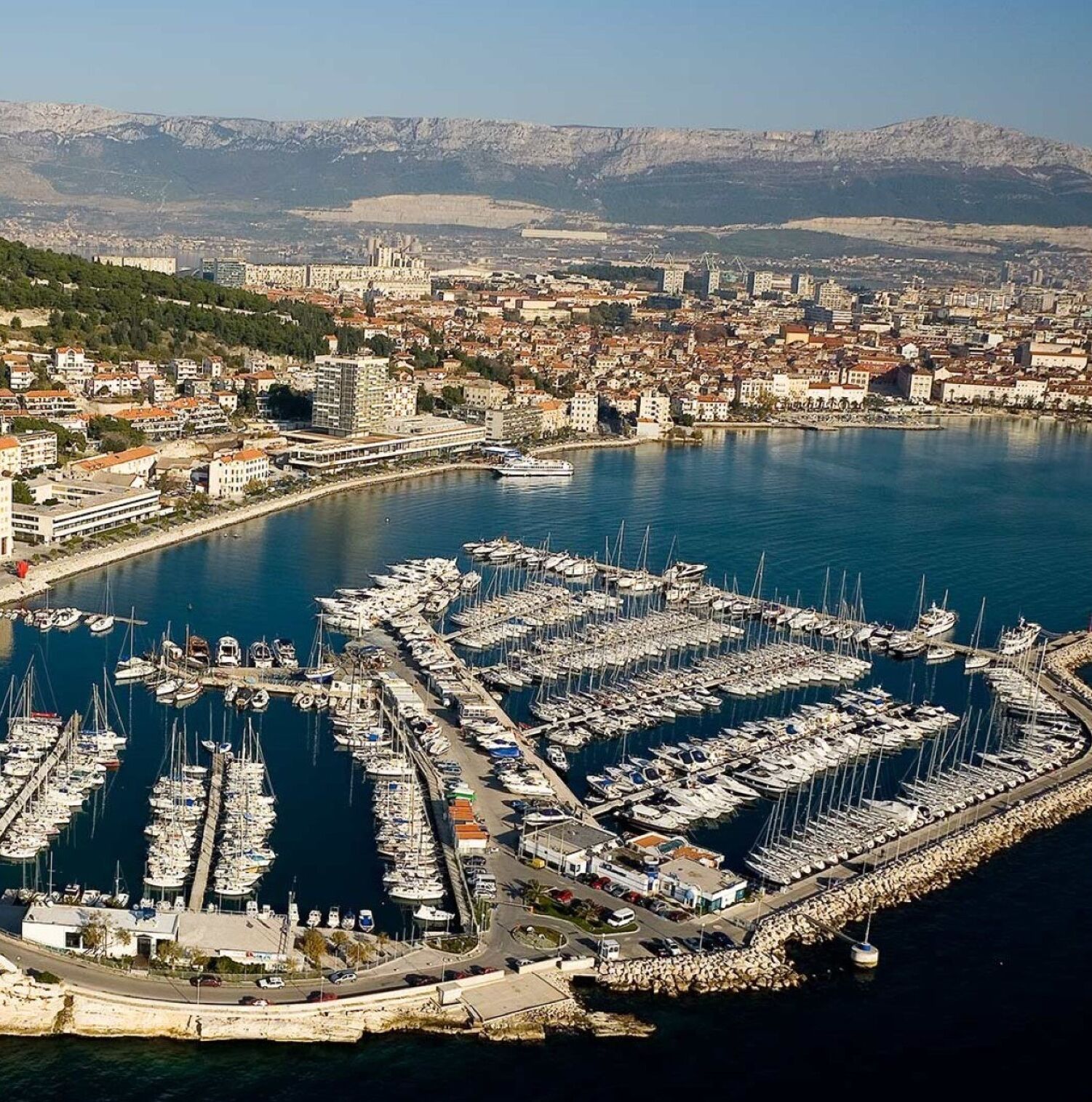 Une vue aérienne d’une marina où sont amarrés de nombreux bateaux, entourée d’un paysage urbain côtier avec des bâtiments et une chaîne de montagnes en arrière-plan.