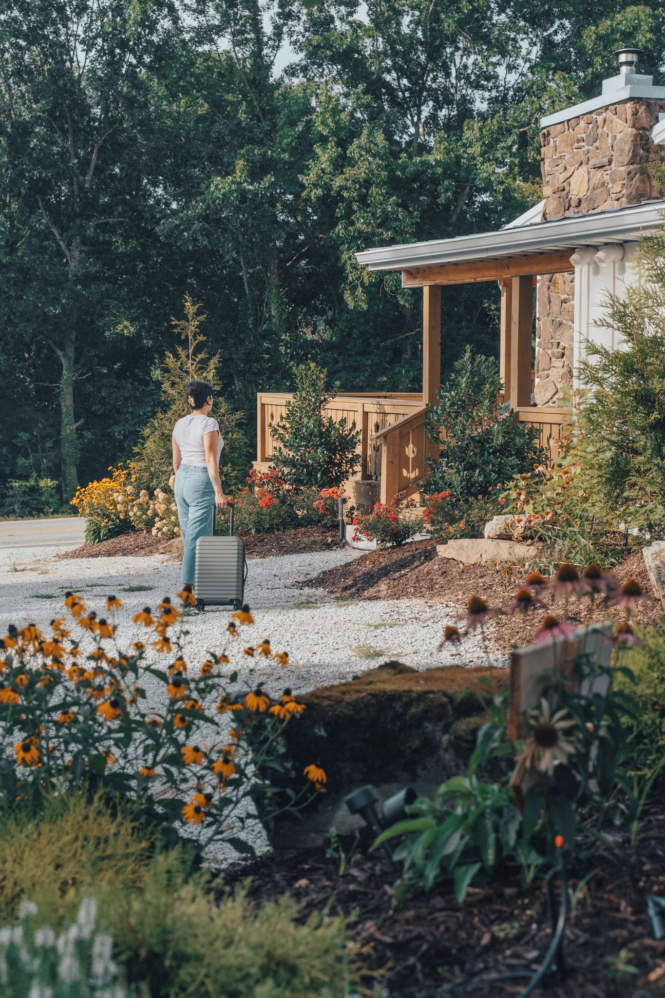A person with a suitcase approaching a stone building with a wooden porch and a garden with various plants and flowers.