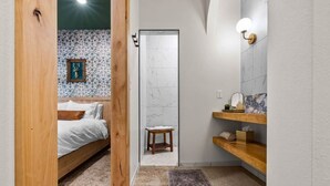Un dormitorio con cama de madera, un taburete pequeño y un baño con pared de mármol y estantes de madera.