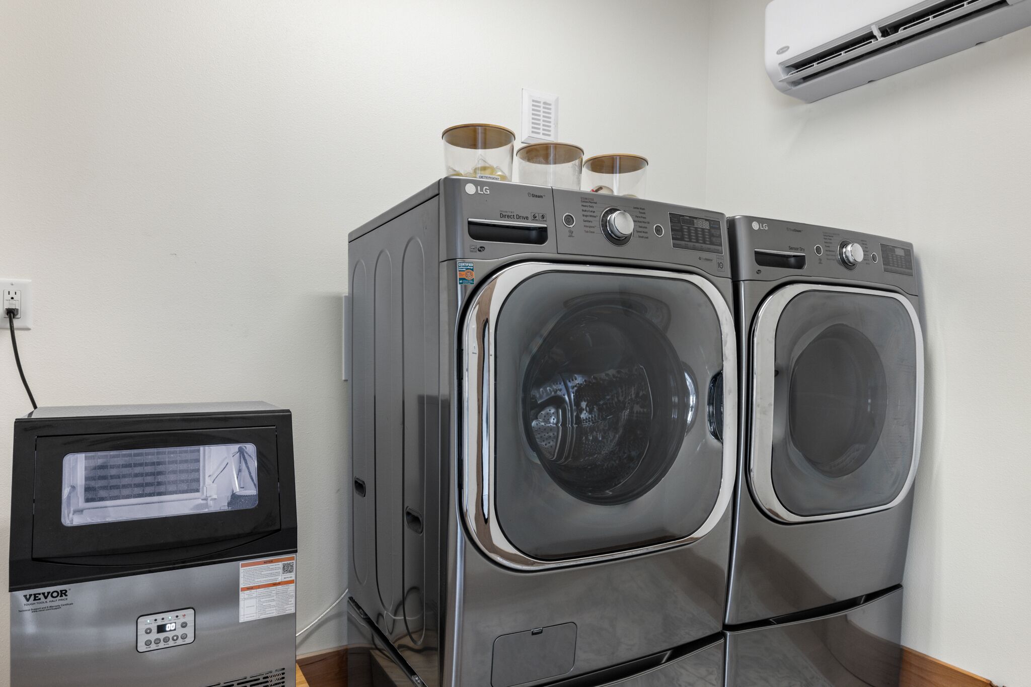 A laundry area with two front-loading LG washers, a VEVOR refrigerator, and an air conditioning unit on the wall.
