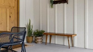 Una habitación con techo y puerta de madera, una silla de metal negro, un banco, un estante con una planta y un cuadro enmarcado.