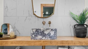 Une salle de bain avec un meuble-lavabo en bois, un évier en marbre, deux appliques murales, un miroir et une plante en pot.