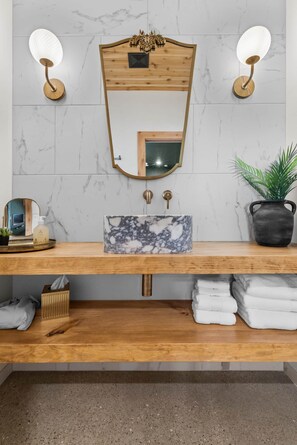 Une salle de bain avec un meuble-lavabo en bois, un évier en marbre, deux appliques murales, un miroir et une plante en pot.