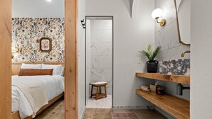A bedroom with a bed, a wooden door, and a bathroom with a sink, mirror, and towel rack.