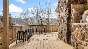 Une terrasse en bois avec des tabourets de bar, un mur en pierre et une vue sur des arbres sans feuilles et des montagnes.