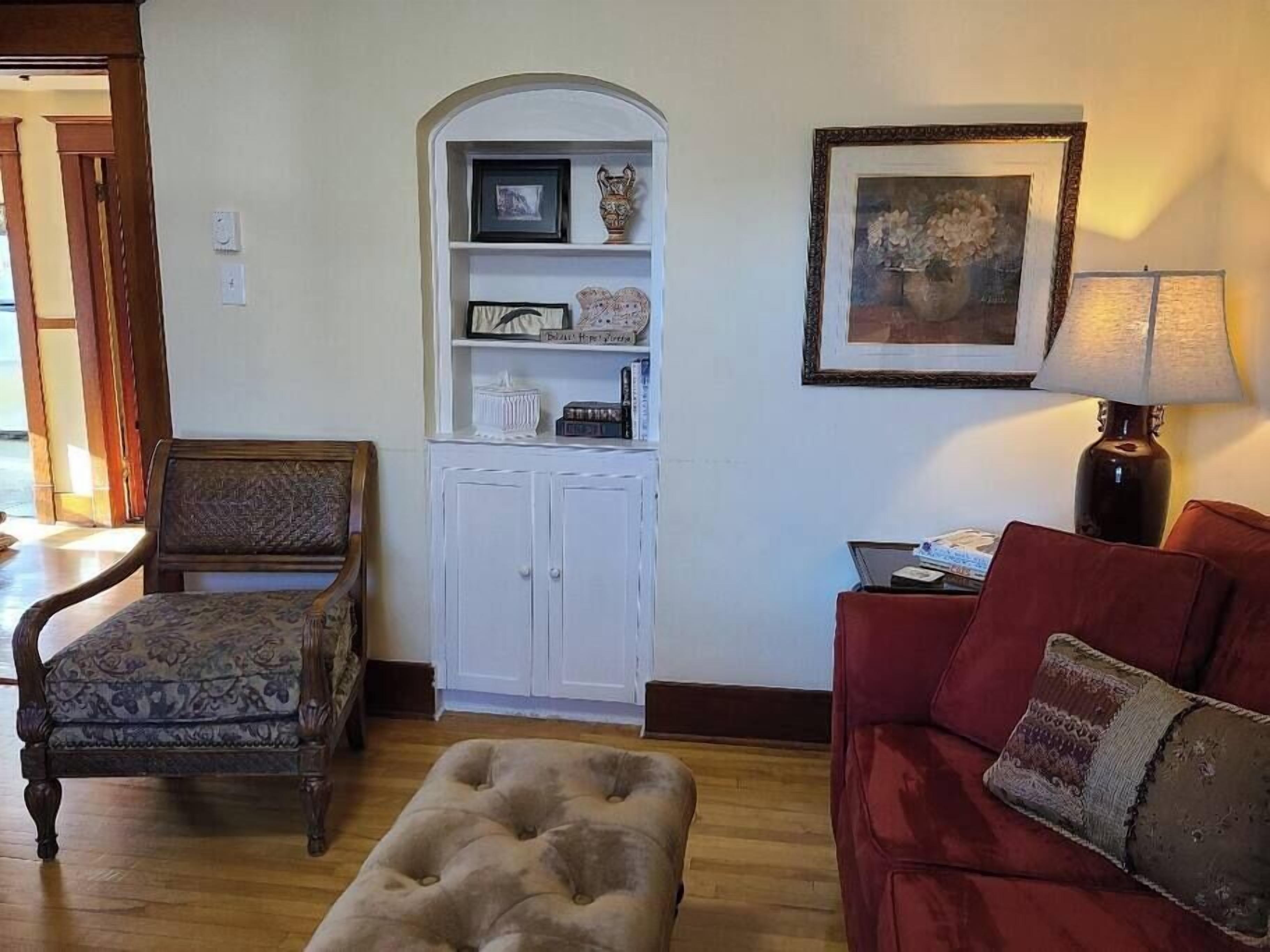 A living room with a wooden chair, a red sofa, a tufted ottoman, a white cabinet with shelves, and a framed picture on the wall.