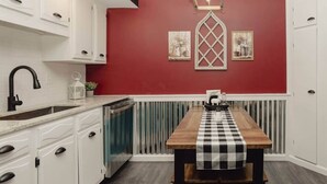 A kitchen with white cabinets, a red accent wall, a checkered table runner, and a window with a decorative grille. - First Floor Getaway in Meadville (West Mead Township)