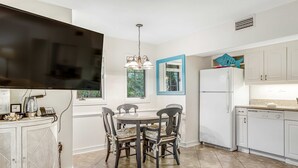 A modern kitchen with white cabinets, a dining table with chairs, a large flat-screen TV, and a blue-framed window. - Charming condo with private beach access, on-site golf, high-speed WiFi & AC (Kiawah Island)