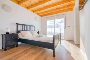Een slaapkamer met een houten plafond, een bed met donker frame, een nachtkastje en een groot raam.