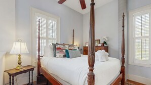 Ein Bett mit Baldachin, mit weißer Bettdecke, ein Nachttisch mit Lampe und ein Fenster mit Jalousien.