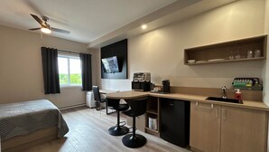 A compact hotel room with a kitchenette, a bed, a desk, and a TV mounted on the wall. - Hôtel St-Michel (Saint-Michel-des-Saints)