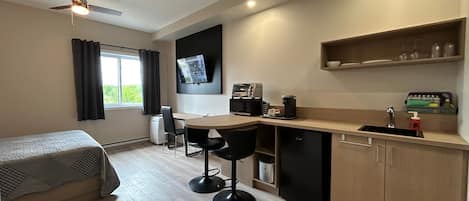 A compact hotel room with a kitchenette, a bed, a desk, and a TV mounted on the wall.