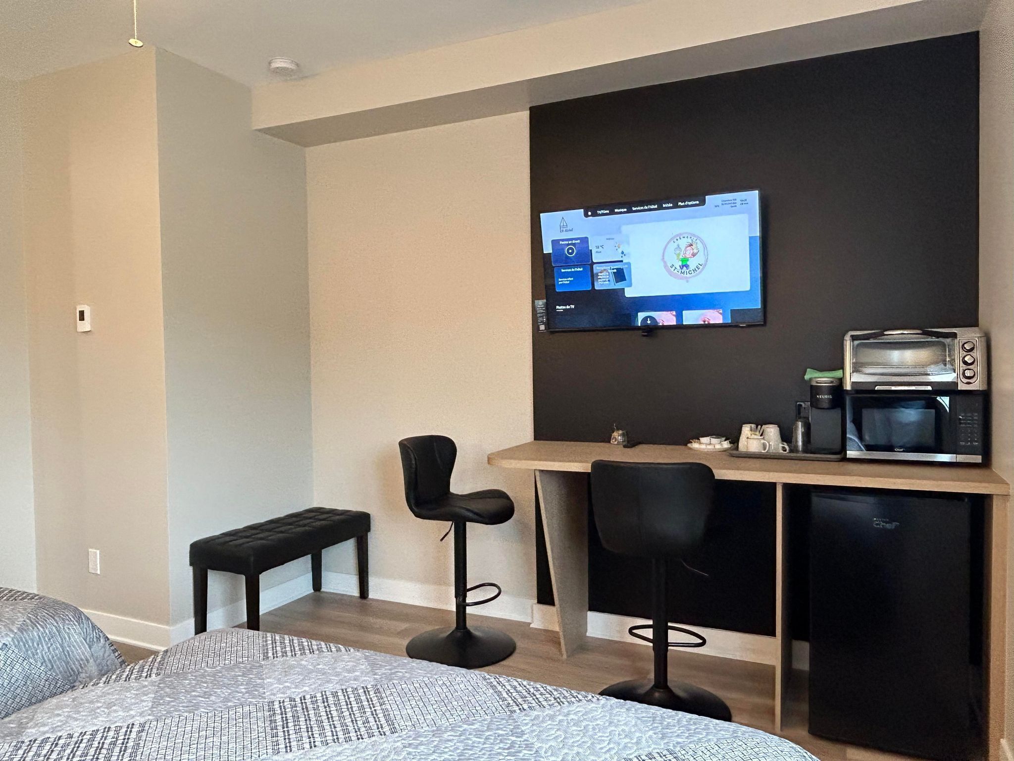 Una habitación de hotel moderna con una cama, un escritorio con una computadora, una cafetera y dos taburetes de bar.
