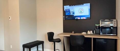 A modern hotel room with a bed, a desk with a computer, a coffee machine, and two bar stools.