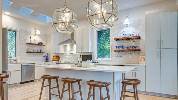 A modern kitchen with a central island, bar stools, pendant lights, and a view of greenery outside.