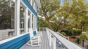 A wooden deck with white rocking chairs and a table, surrounded by trees and a railing.