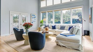 A modern living room with a sofa set, a coffee table, and two armchairs. There are large windows providing natural light and a view of greenery outside.
