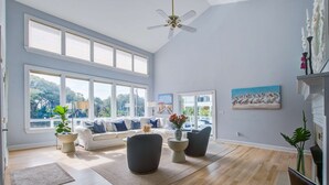 A spacious living room with large windows, a ceiling fan, and a white fireplace.
