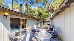 Un patio techado al aire libre con una mesa redonda y sillas, una puerta de vidrio que da a una habitación y una terraza de madera.