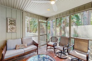 A covered porch with a wooden ceiling, wicker furniture, and large windows offering views of trees and a fence. - Cabin in the maritime forest with pool and golf on-site - walk to the beach (Kiawah Island)