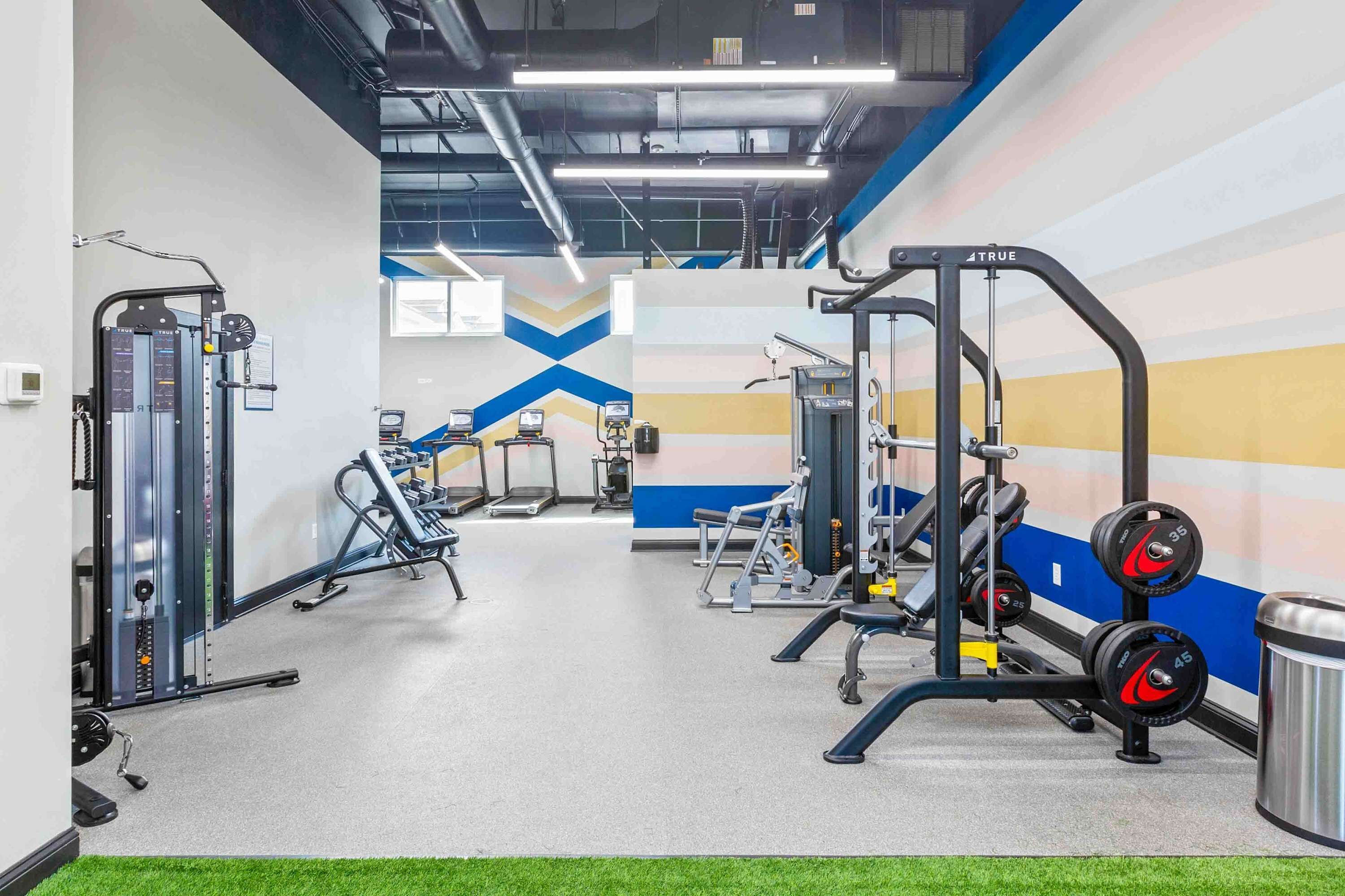 Un gimnasio con diferentes máquinas de ejercicio, una pared con un diseño geométrico y un bote de basura.
