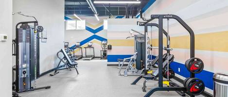 Un gimnasio con diferentes máquinas de ejercicio, una pared con un diseño geométrico y un bote de basura.