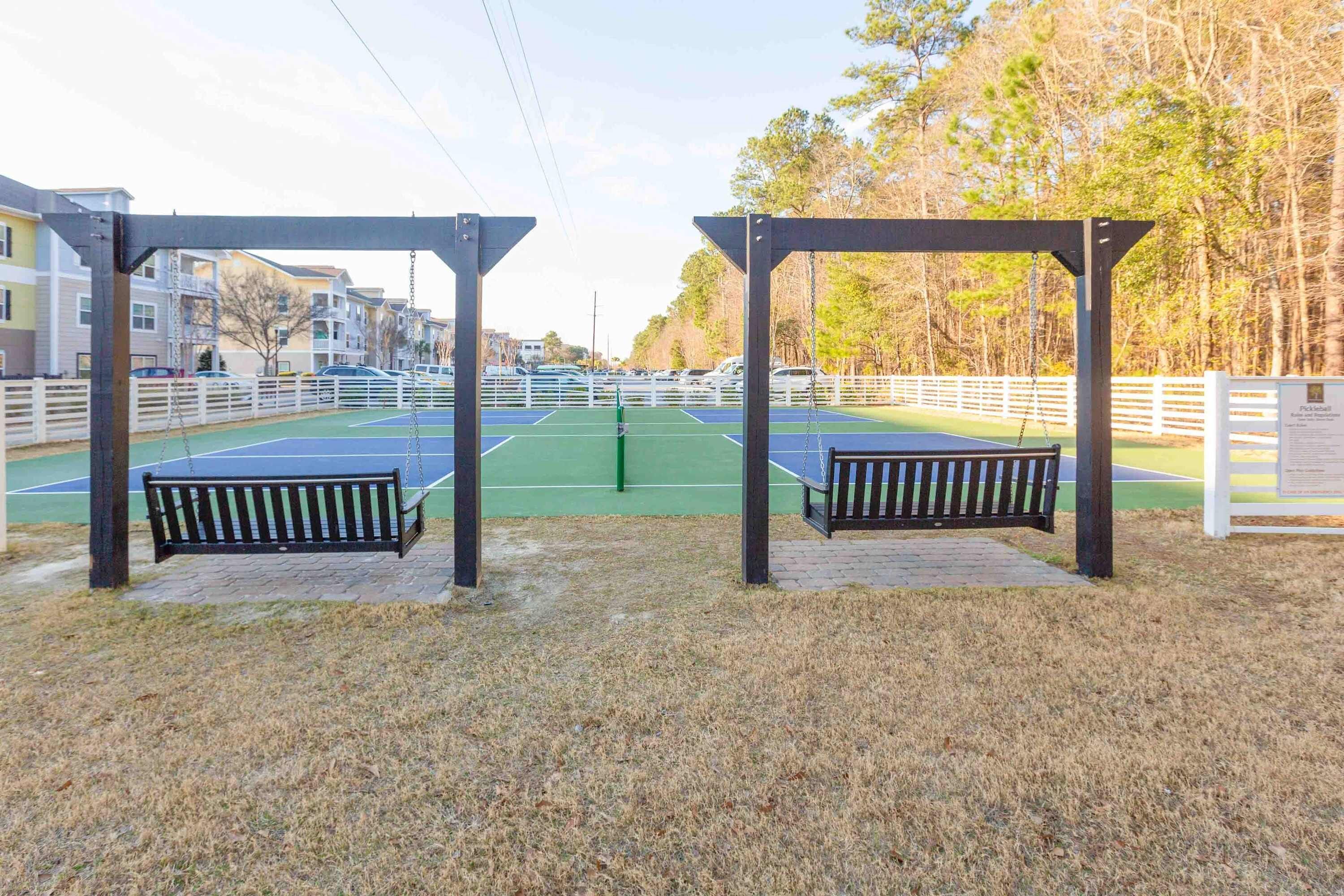 Un court de tennis avec deux bancs placés sous des pergolas, entouré d’une clôture blanche et d’arbres.