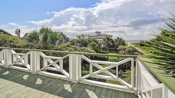 Um deck de madeira com guarda-corpo branco, com vista para um jardim ajardinado e uma casa grande com fachada branca e telhado marrom.