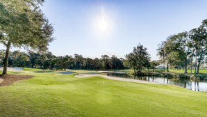 A well-maintained golf course with a pond, trees, and a clear sky.