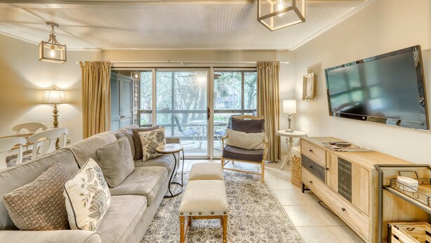 A spacious living room with a sofa, ottoman, and armchair, a television, and a large window with curtains.