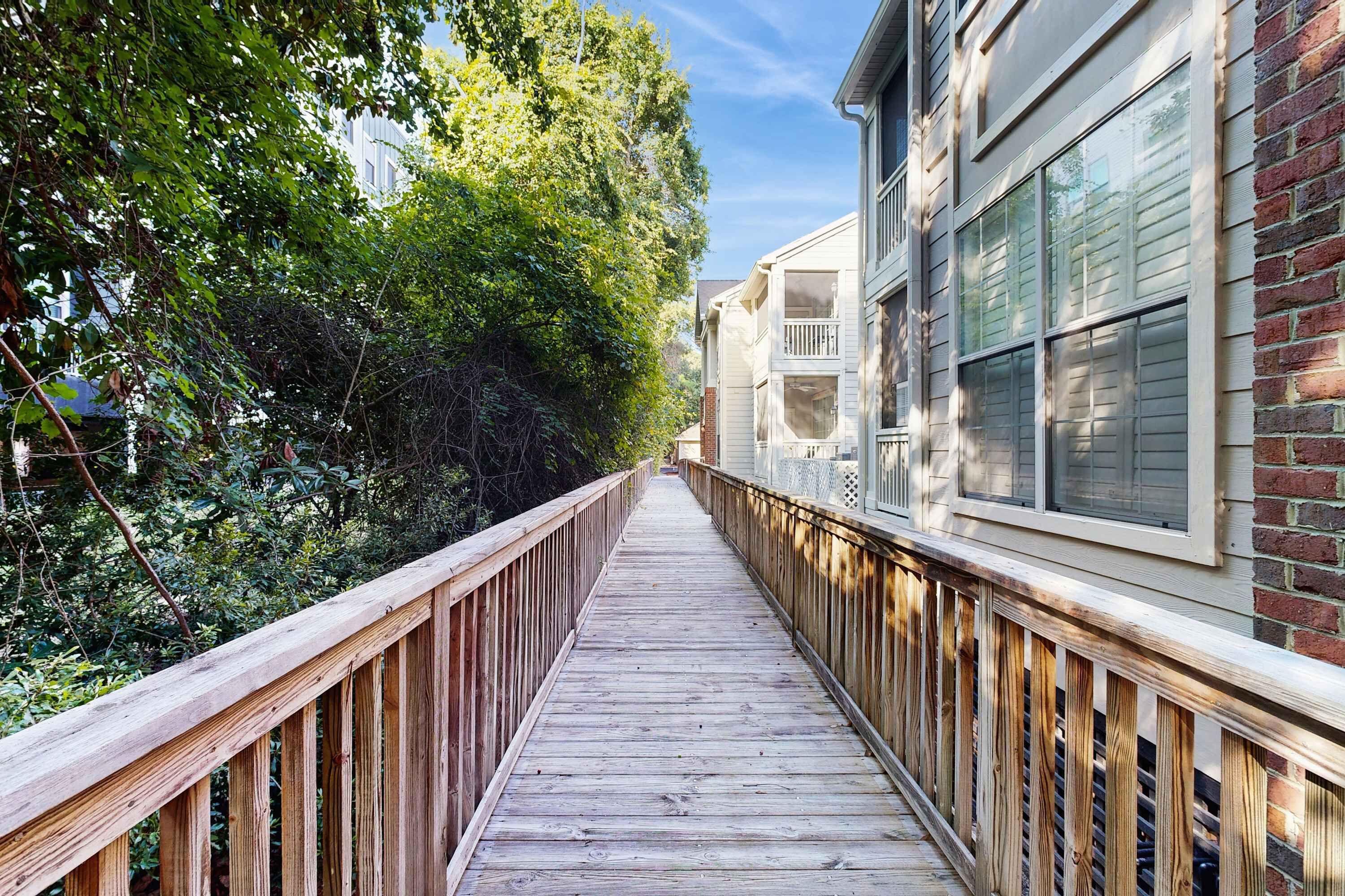 Une terrasse en bois dotée d’une rambarde, entourée d’une végétation luxuriante et bordée de bâtiments adjacents.