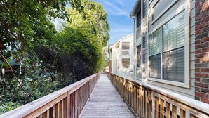 A wooden deck with a railing, surrounded by dense foliage and adjacent buildings.