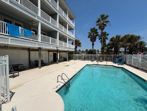 A pool area with a clear blue pool, lounge chairs, and a building with balconies.