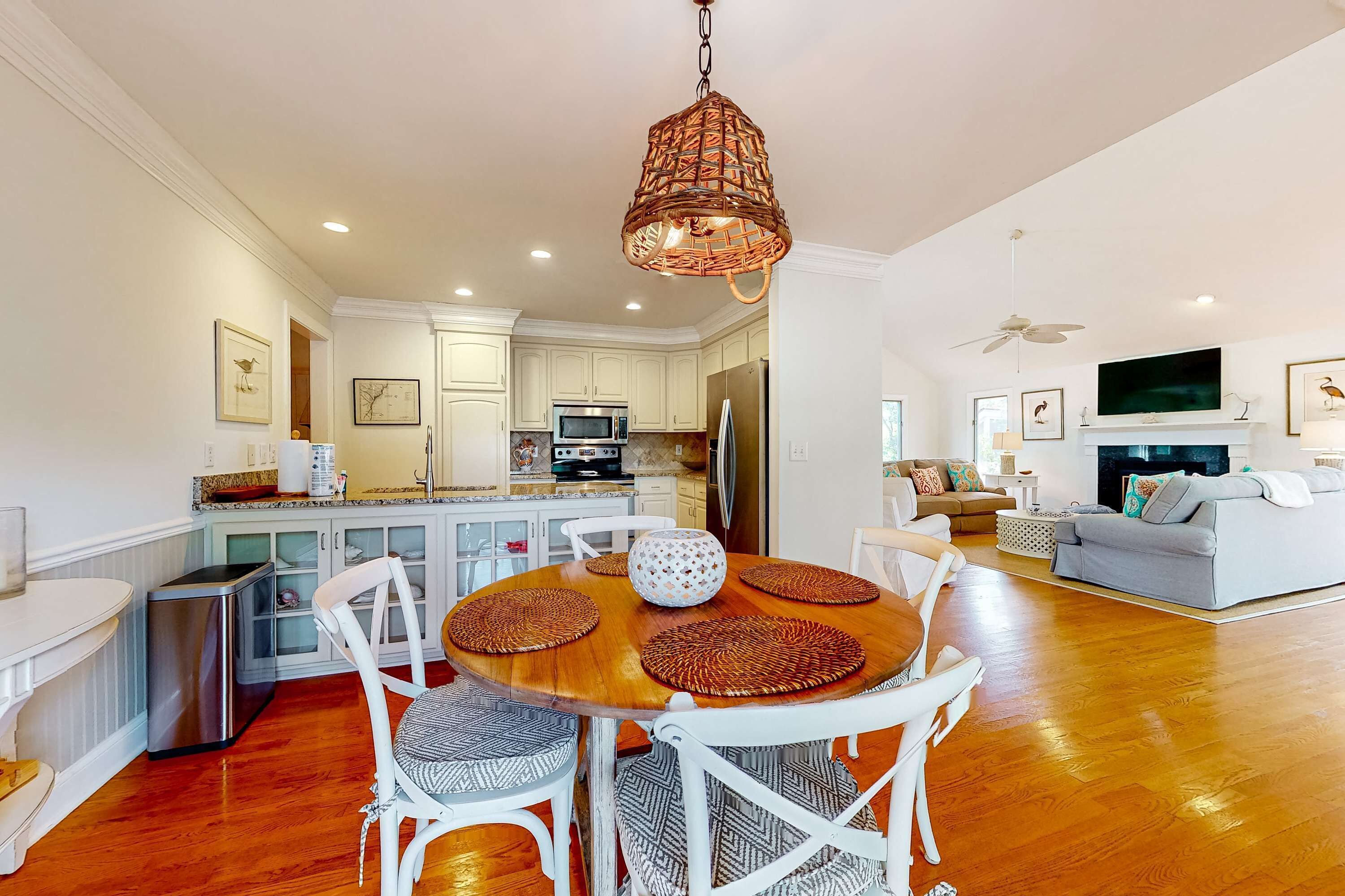 Uma cozinha moderna com armários brancos, uma sala de jantar com mesa redonda e cadeiras brancas, e uma sala de estar com sofá e televisão.