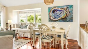 Sala de estar luminosa com sofá, mesa de jantar e cadeiras. Há um quadro na parede e um lustre.