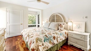 Quarto com cama, mesa de cabeceira, cômoda, ventoinha no teto e janela.