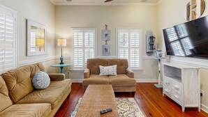 A living room with a beige sofa, a brown armchair, a coffee table, and a TV on a white cabinet. - North Central, 30-Day Rental Home w/Double Porches, Garden Patio, and Free WiFi (Charleston)