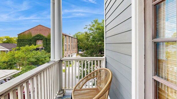 A balcony with a wicker chair, white railings, and a view of neighboring buildings and trees. - North Central, 30-Day Rental Home w/Double Porches, Garden Patio, and Free WiFi (Charleston)
