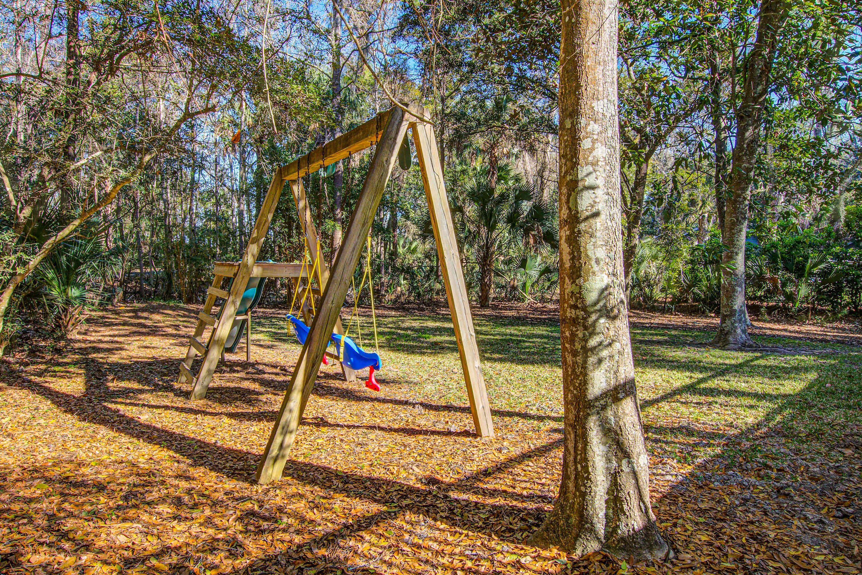 A wooden swing set in a wooded park area with trees and fallen leaves on the ground.