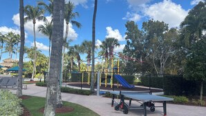 Un terrain de jeux équipé d’un toboggan et de structures d’escalade, entouré de palmiers et bordé d’un court de tennis.