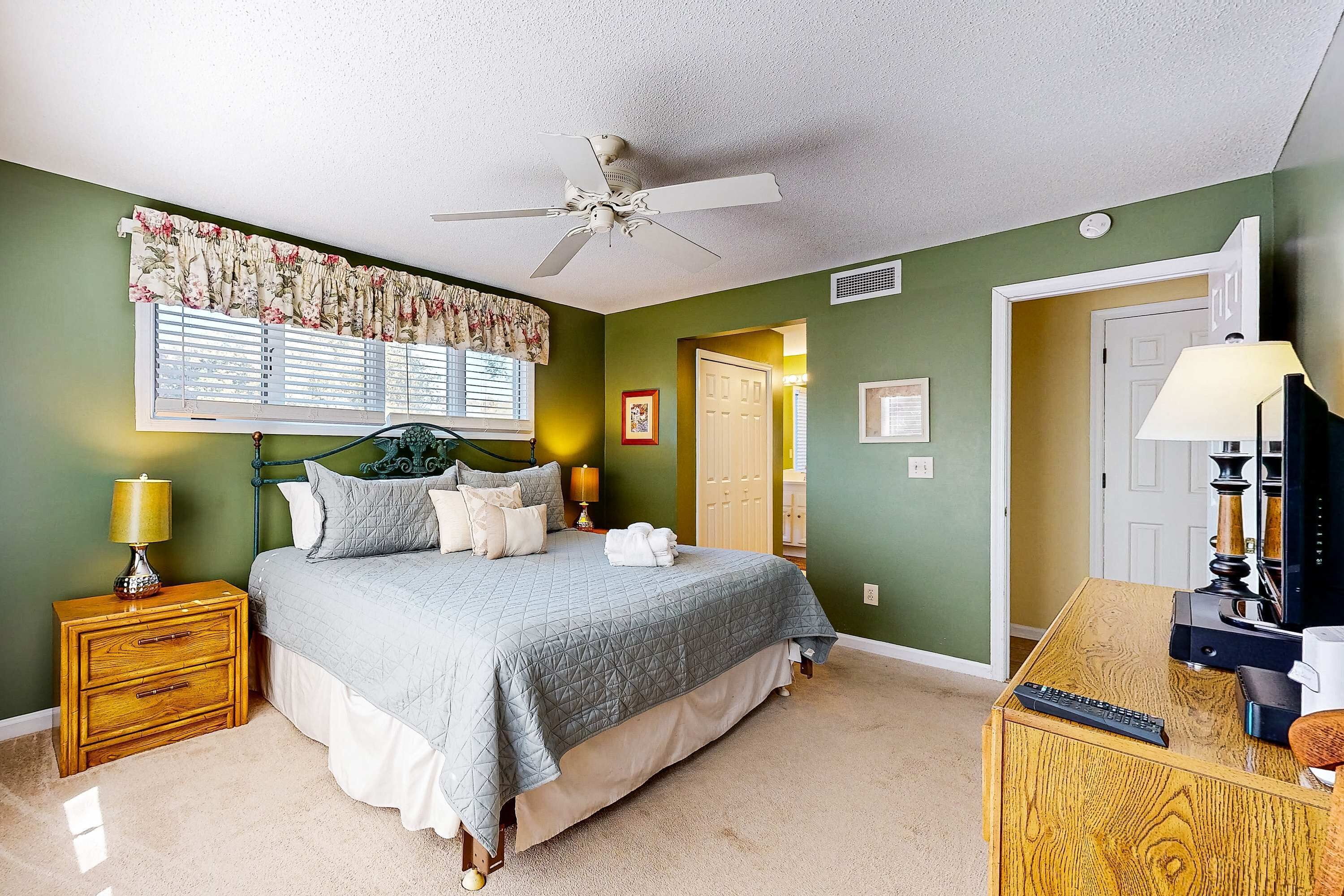 A bedroom with a bed, nightstand, lamp, and a desk with a lamp and a remote control.