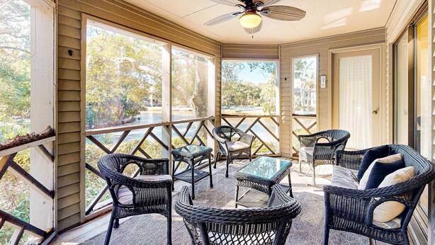 A covered outdoor seating area with wicker furniture, a ceiling fan, and large windows offering views of the surrounding trees.