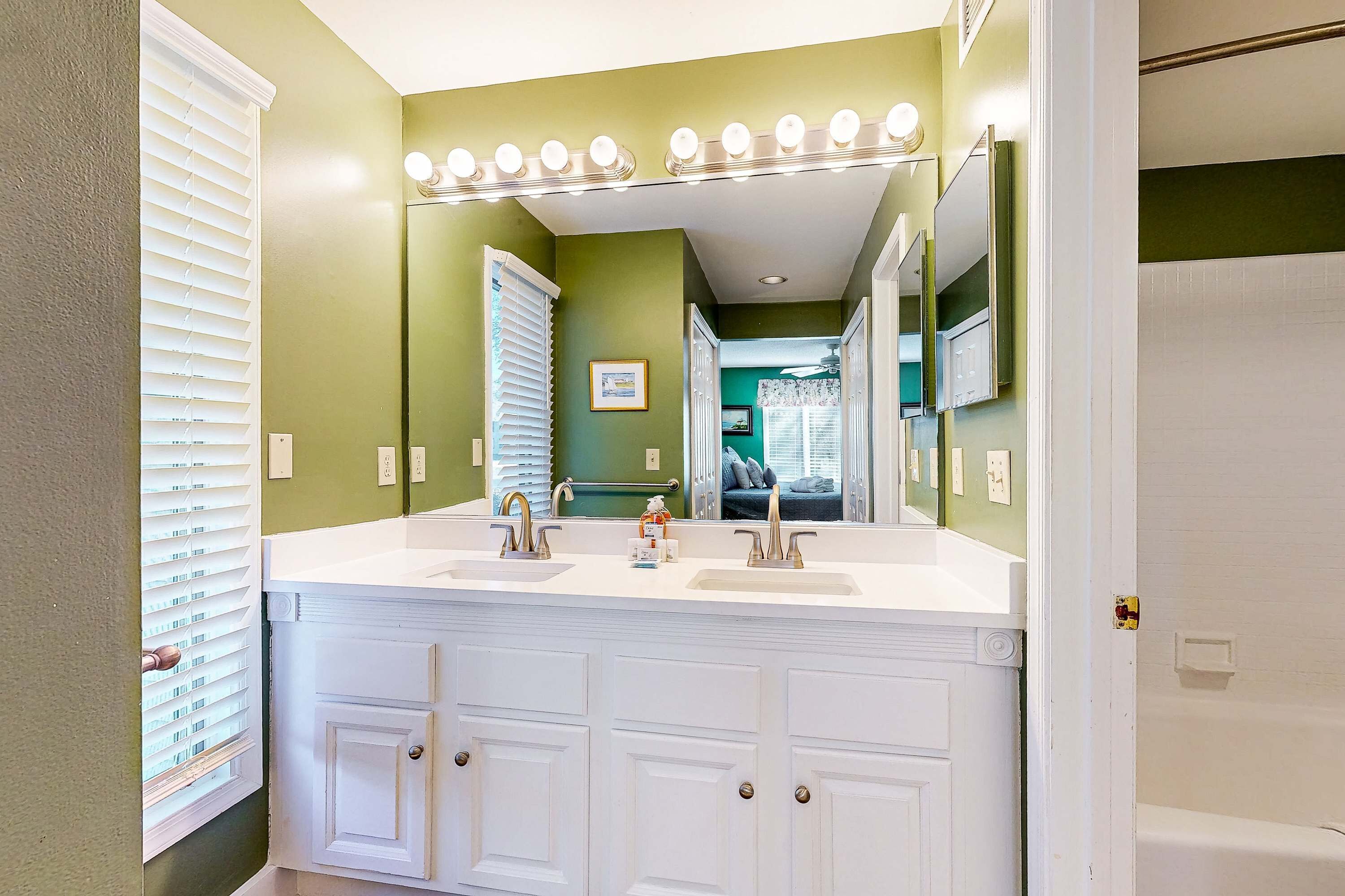 A bathroom with a double sink vanity, a large mirror with lights, and a view into the adjacent room.