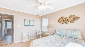 Um quarto com uma cama, um ventilador de teto, uma pintura de peixes na parede, uma cadeira, uma mesa pequena e uma porta que dá para outro cômodo.