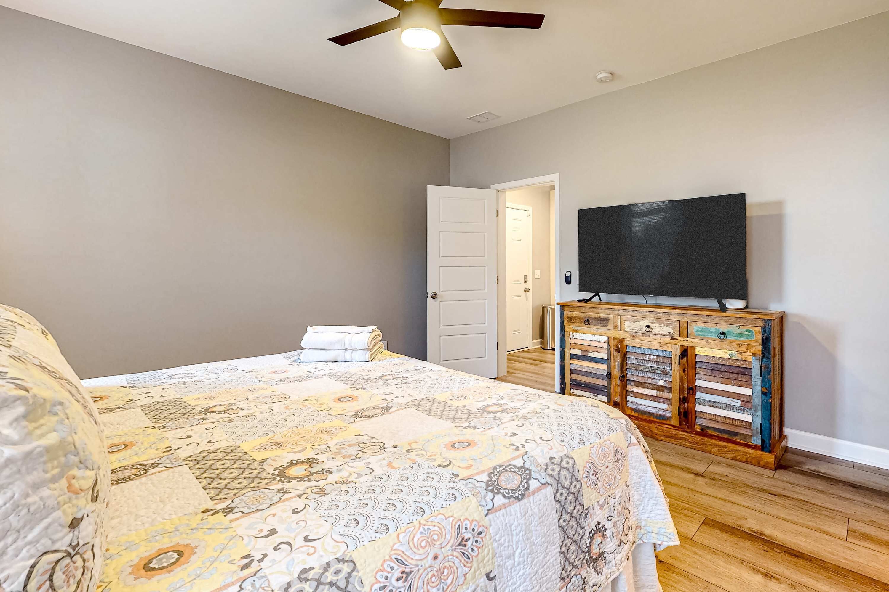 Un dormitorio con una cama grande, un ventilador de techo, un televisor sobre un mueble de madera y una puerta que da a otra habitación.