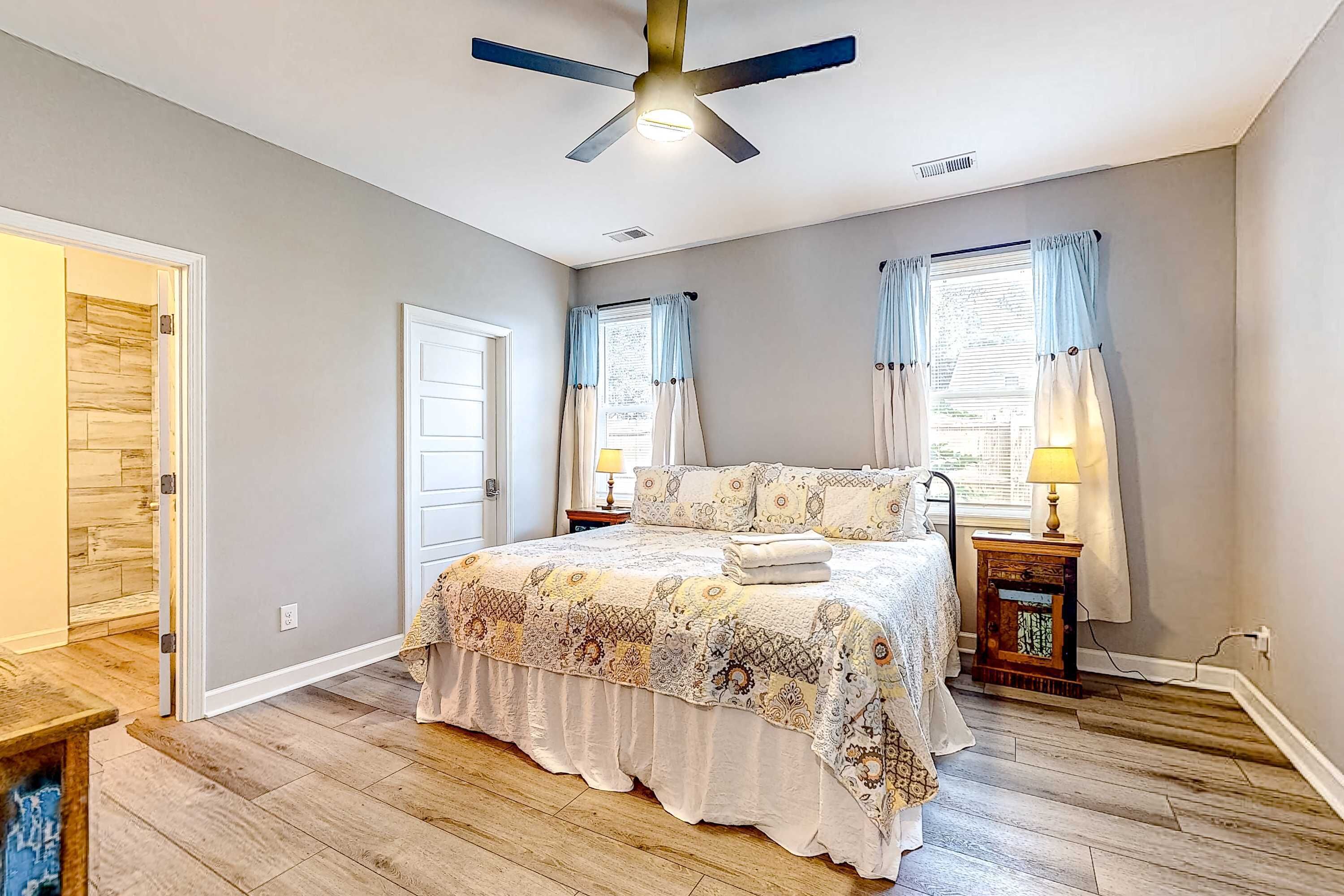 A bedroom with a bed, a ceiling fan, two windows with curtains, a bedside table with a lamp, and a wooden floor.