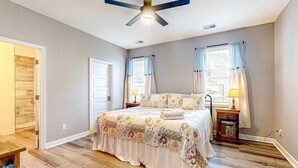 Un dormitorio con cama, ventilador de techo, dos ventanas con cortinas, mesita de noche con lámpara y suelo de madera.