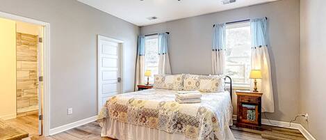 A bedroom with a bed, a ceiling fan, two windows with curtains, a bedside table with a lamp, and a wooden floor.