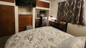 Une chambre avec un lit, un téléviseur fixé au mur, un bureau avec une chaise, un four à micro-ondes et une petite poubelle.